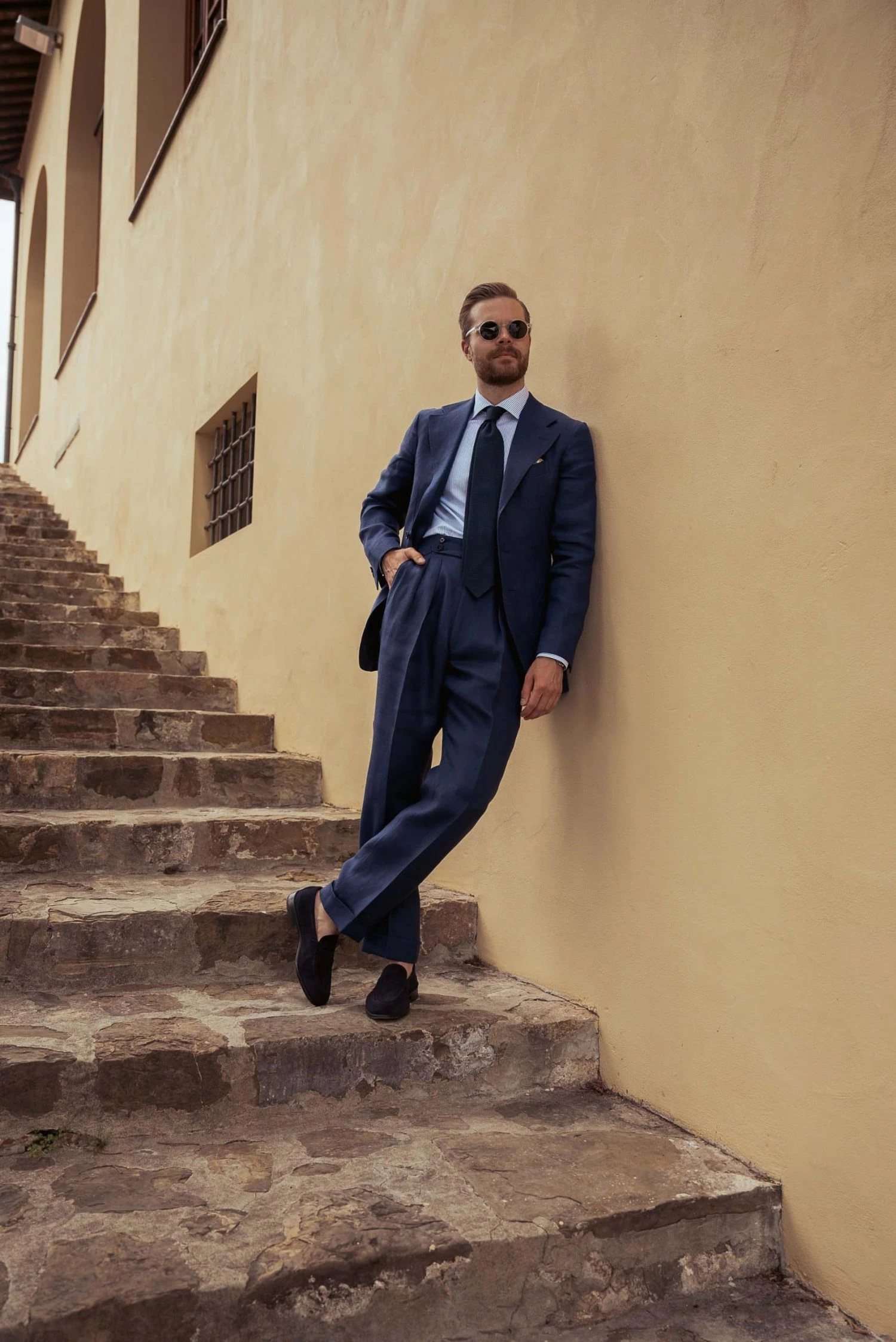 distance shot Johan Wikstrøm on old stoneslab stairs leaning against a wall, wearing sunglasses and a custom navy blue linen suit at Pitti Uomo