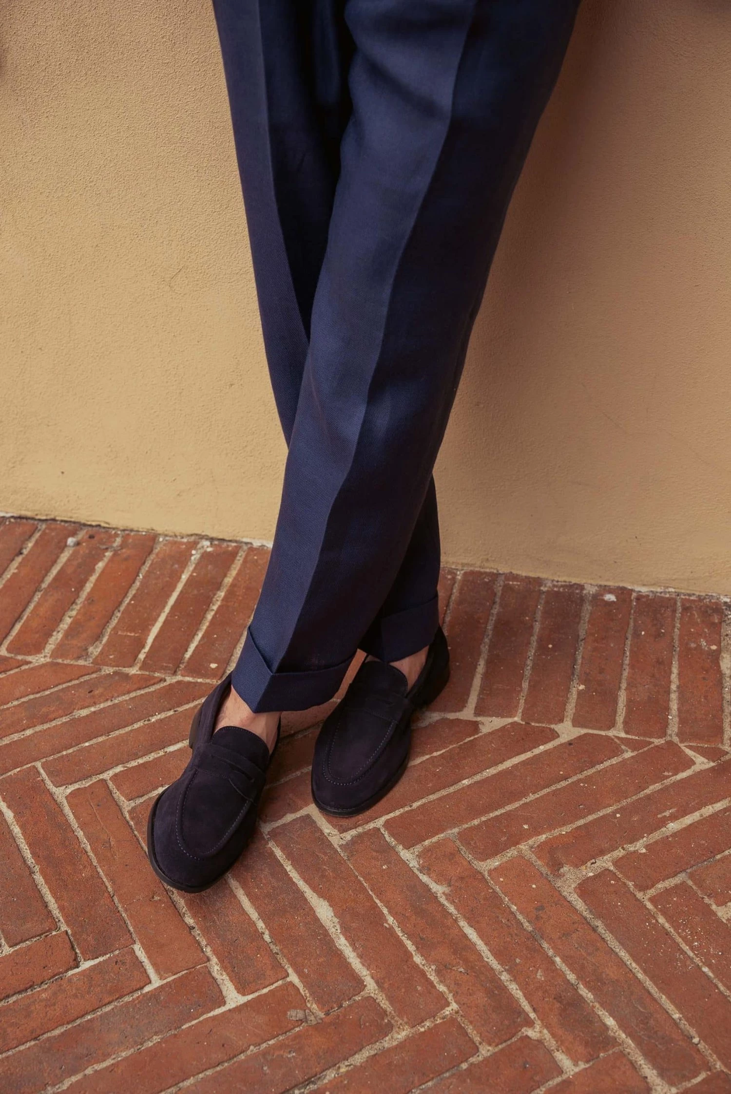 Legs crossed, in navy blue suit trouser legs and blue loafers, standing on herringbone brick floor