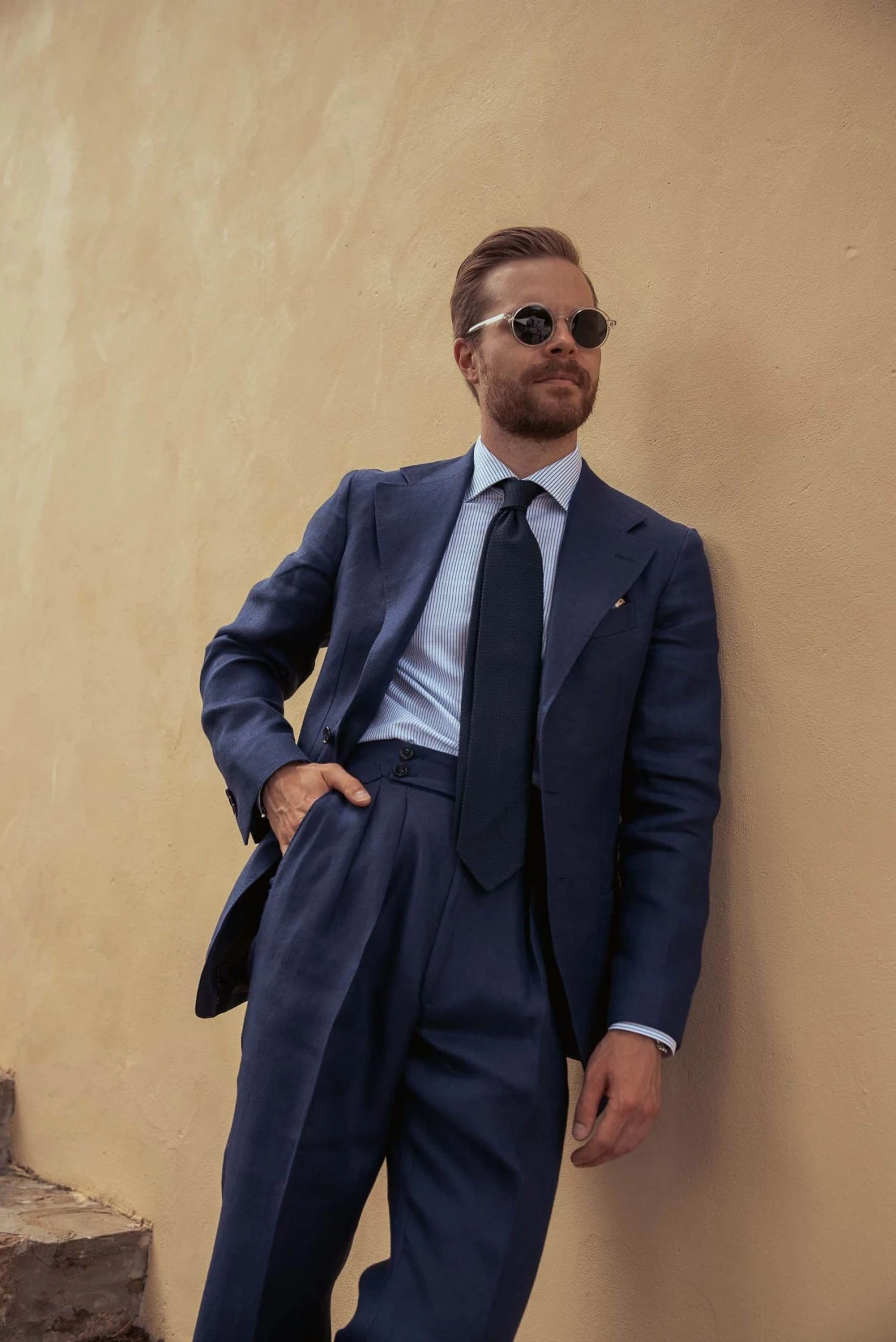 Johan Wikstrøm leaning against a wall, wearing sunglasses and a custom navy blue linen suit at Pitti Uomo