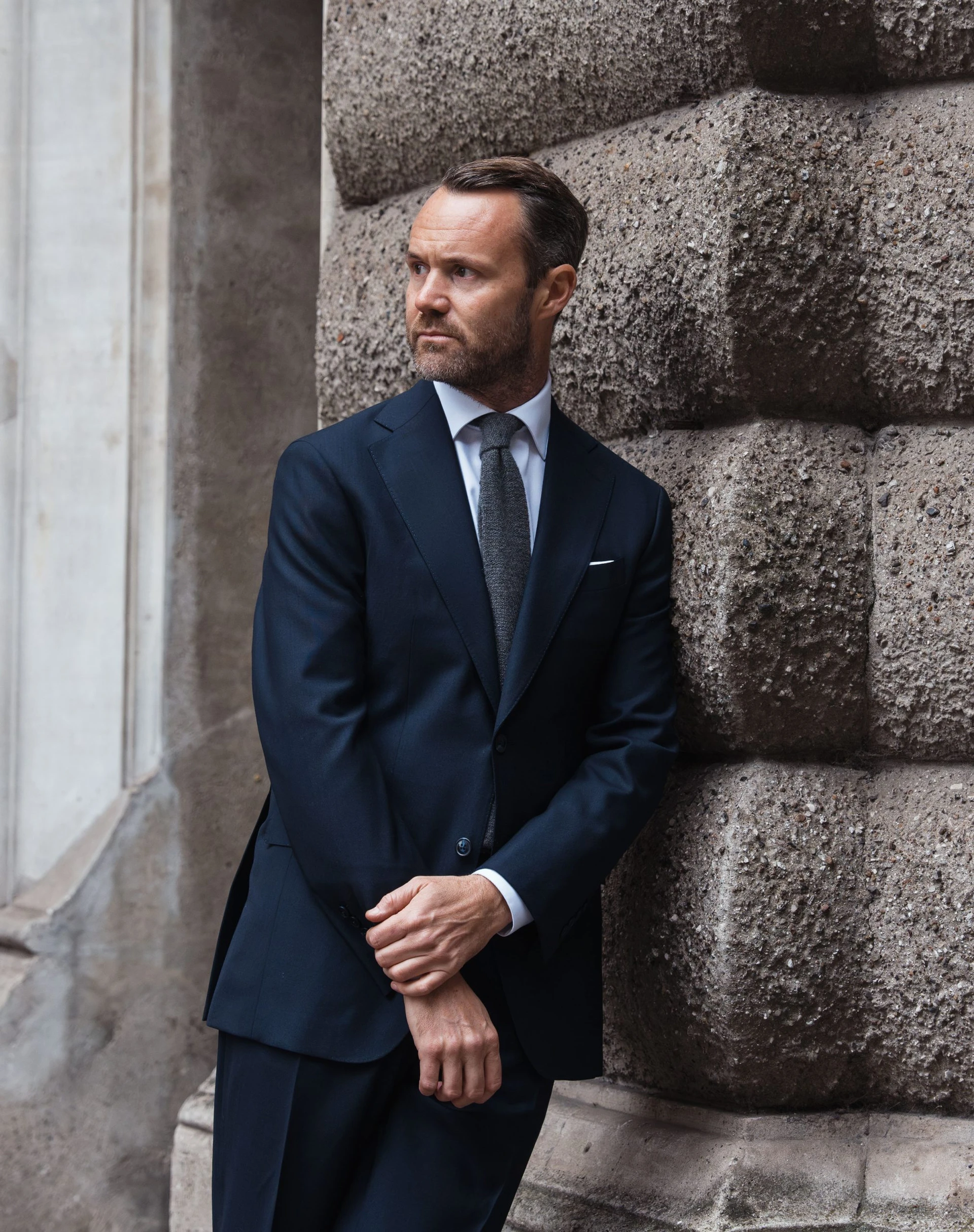 man leans against stone wall in a navy dark blue suit in twistair fabric