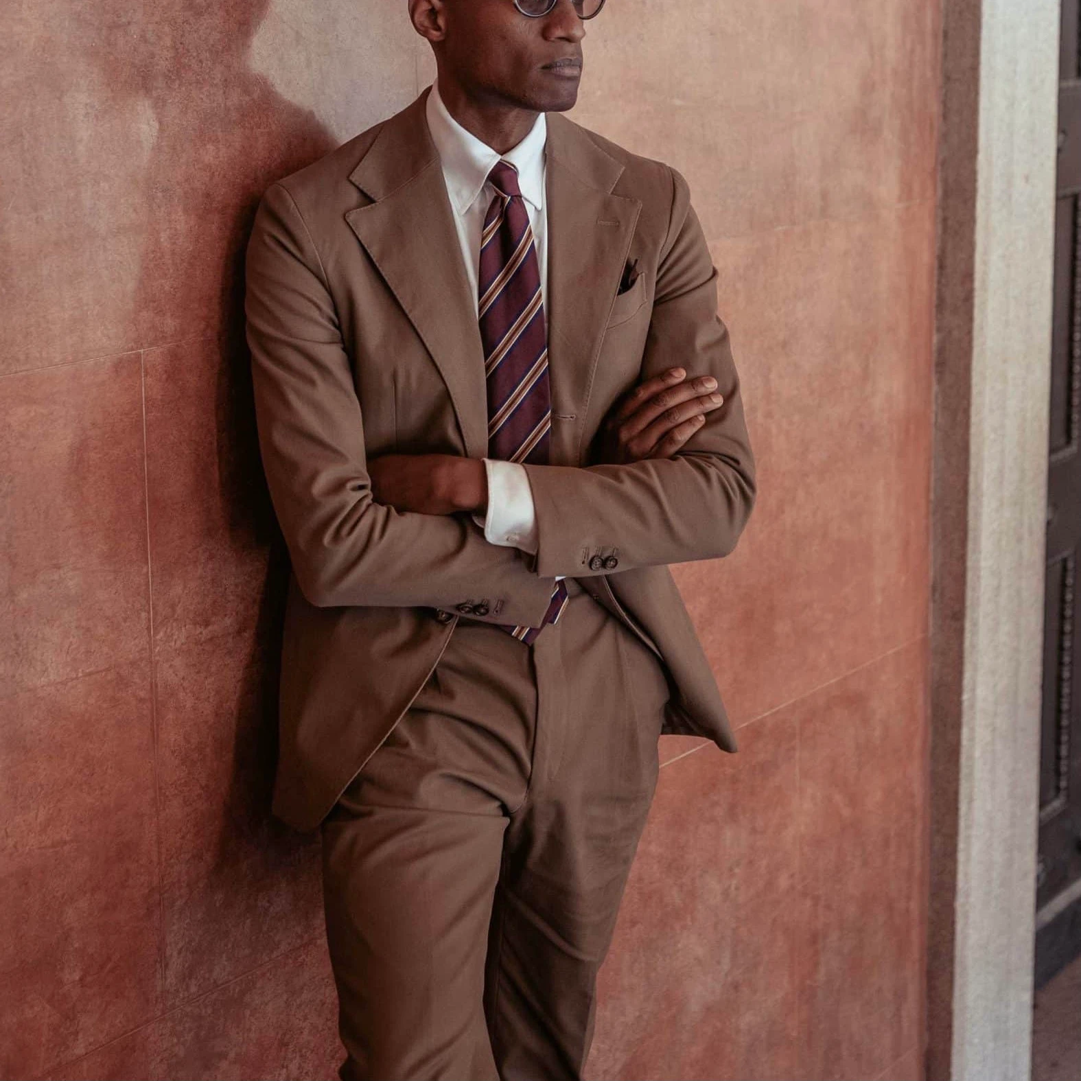 Man leaning against wall, standing with his arms crossed. He's wearing a taupe cotton suit, a white shirt and a striped burgundy tie